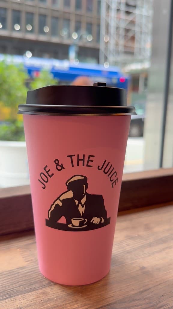 A pink Joe & The Juice cup with a bold black logo, photographed against a blurred city street.