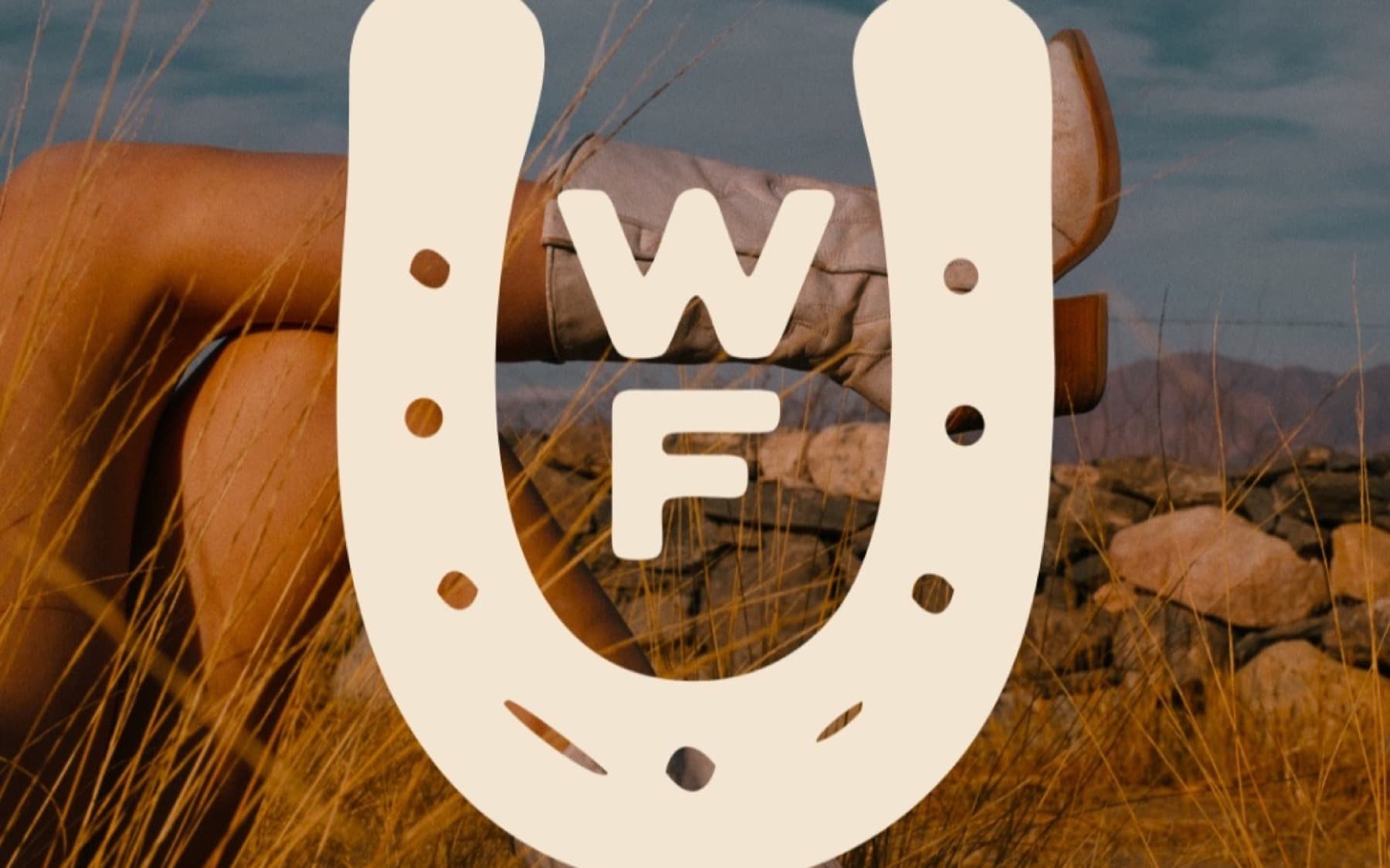 Wild Form horseshoe mark over western boots in tall grass with a stone wall and open sky behind them.